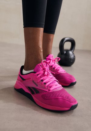 Fødder iført lyserøde Reebok-sneakers og sorte leggings, stående på en lys gulv med en sort kettlebell i baggrunden.
