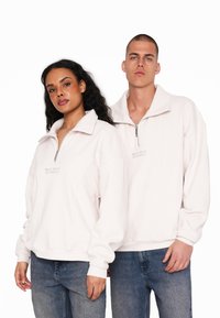 Lys pink zip-up pullover lavet af blødt stof, med høj krave, broderet logo og tætsiddende manchetærmer. Stylet med denim.
