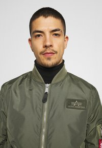 Giacca bomber verde oliva con zip argentata, colletto a coste e etichetta a patch. Presenta tasche laterali e una texture di tessuto liscia e lucida.
