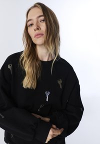 Czarna bluza z długim rękawem, zdobiona wzorami klucza i dziurki od klucza z metalicznymi akcentami. Luźny krój, z teksturowanym materiałem.