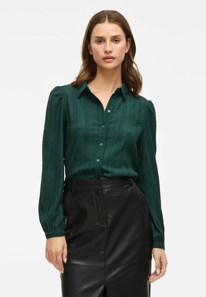 Donkergroene blouse met een gestructureerd patroon, knoopsluiting aan de voorkant en lange pofmouwen, gecombineerd met een zwarte leren rok met een split.
