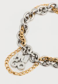 Βραχιόλι αλυσίδα σε ασημί και χρυσό τόνο με μεγάλα συνδετικά κρίκους, διακοσμημένο με μεταλλικό γούρι "HUGO". Λείο υφή με αντίθετες επιφάνειες φινιρίσματος.