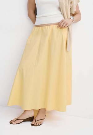 Donna che indossa una gonna a linea A giallo chiaro, top bianco senza maniche, maglione beige drappeggiato sulla spalla e sandali con zeppa marroni.