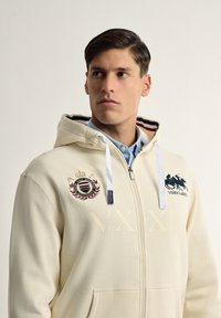 Sudadera con cremallera en color crema que presenta un parche con logo, detalles bordados, bolsillo tipo canguro y forro de rayas en contraste en la capucha.