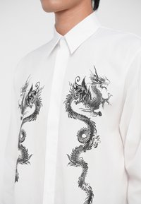 Cămașă albă cu nasturi, prezentând broderie de dragon negru care se învârte pe ambele laturi, evidențiind detalii intricate și o textură fină a fabricii.