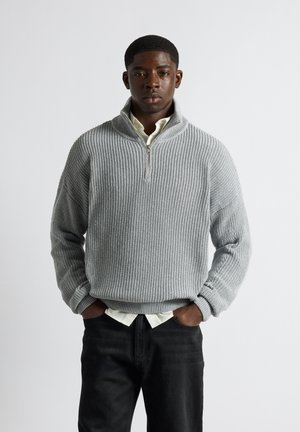 Jeune homme les mains dans les poches portant un pull gris clair côtelé à demi-fermeture éclair sur une chemise blanche et un pantalon noir, debout devant un fond uni.