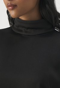 Maglia nera a collo alto finto realizzata in tessuto liscio e leggero, caratterizzata da una silhouette aderente e un design minimalista con una leggera lucentezza.