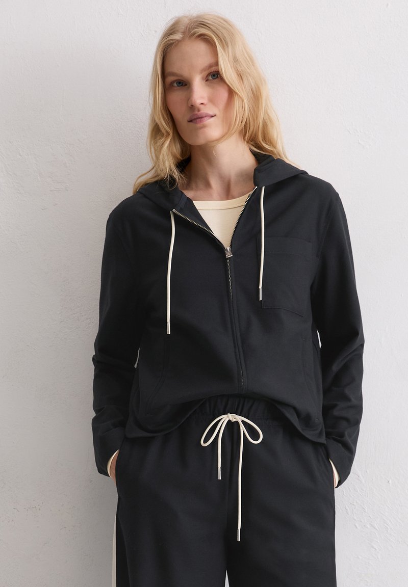 Schwarzer Hoodie mit durchgehendem Reißverschluss, Kordelzug, einer Brusttasche und lockerem Schnitt. Weiches Material mit glatter Textur, kombiniert mit passenden Hosen.