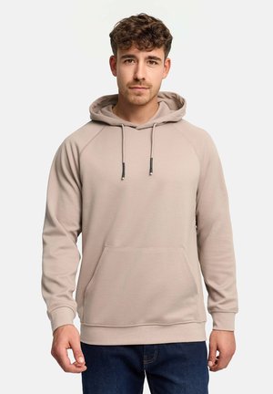 Jonge man met kort kroeshaar, gekleed in een beige hoodie met een voorkantzak en donkerblauwe spijkerbroek, staand tegen een effen lichte achtergrond.