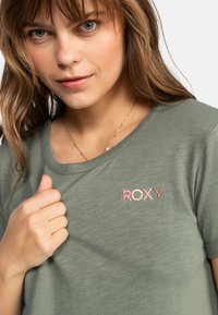 Zelené tričko s krátkým rukávem, kulatým výstřihem a vyšitým logem Roxy v růžové a oranžové barvě na hrudi, vyrobené z měkké tkaniny.