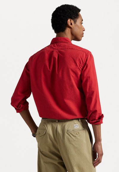 Punane pika varrukaga särk, millel on nööbiga krae ja üleskeeratud varrukad, kombineeritud heledate beezide pükstega, millel on tagataskud ja sildi detail.