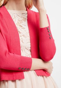 Korallfärgad cardigan i ett slätt stickat tyg, kort design, med ribbade ärmslut, tre svarta knappar och en rundad öppen framsida.