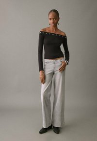 Crna off-shoulder bluza s dugim rukavima i metalnim okomitim rupama, u kombinaciji sa svijetlosivim širokim trapericama i crnim čizmama do gležnja na neutralnoj pozadini.