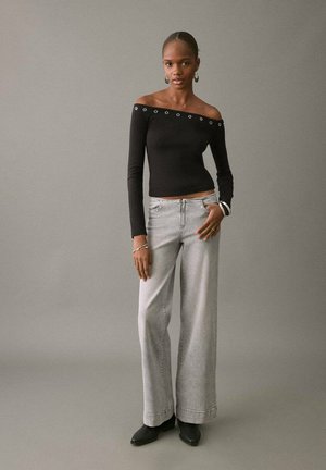 Schwarzes Off-Shoulder Langarmoberteil mit metallischen Ösen, kombiniert mit hellgrauen, weiten Jeans und schwarzen Ankle Boots vor einem neutralen Hintergrund.