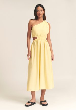 Žluté šaty na jedno rameno s elastickým pasem, bočním výstřihem a splývavou sukní. Texturovaná látka, doplněné černými sandály na plochém podpatku.