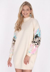 Strickkleid in Creme mit langen Ärmeln, die mit mehrfarbigen Pailletten geschmückt sind, und einem hohen Kragen. Weicher Stoff und lockere Passform.