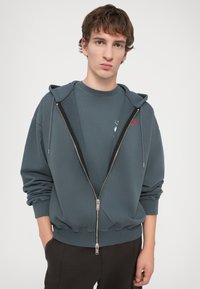 Sweatshirt à capuche zippé en tissu bleu-gris muted, avec des poignets côtelés, une fermeture éclair à l'avant et un petit logo sur la poitrine.