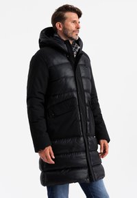 Schwarze Pufferjacke mit Kapuze, die eine Mischung aus glatten und gesteppten Texturen, Druckknöpfen und zwei Fronttaschen aufweist.