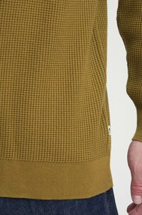 Zielony sweter o strukturalnej teksturze z wzorem w rybkę, z kontrastowymi mankietami i dołem. Szew boczny zawiera małą metkę. Nosi się go na ciemnym dżinsie.
