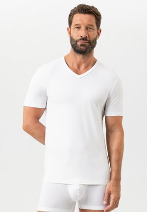 Uomo con barba che indossa una maglietta a V bianca semplice e boxer bianchi, in piedi contro uno sfondo chiaro uniforme.