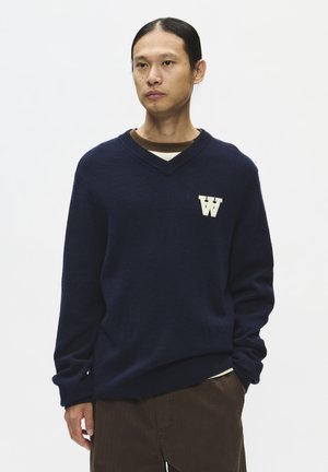 Homme aux cheveux foncés attachés porte un pull bleu foncé à col en V avec un logo blanc en forme de W, superposé à une chemise marron et un pantalon marron sur fond blanc.