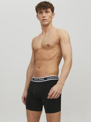 Schwarze Baumwoll-Boxershorts mit einem weißen Logo-Bund, enger Passform und glatter Textur. Das Modell steht in einer neutralen Pose und ohne Hemd.