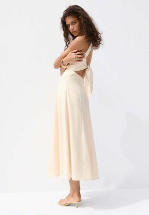 Femme aux cheveux bouclés portant une robe crème sans manches avec un dos ouvert et un détail noué, debout en sandales à talons beiges sur fond blanc.