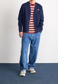 Man som bär en marinblå Nike-kardigan över en röd och vit randig skjorta, lösa blåjeans och beige sneakers, stående mot en vit vägg.