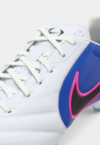 Λευκό ποδοσφαιρικό παπούτσι με μπλε και μαύρες λεπτομέρειες. Διαθέτει ροζ λογότυπο Nike, υφή από δέρμα στην επάνω επιφάνεια και επίπεδα λευκά κορδόνια.