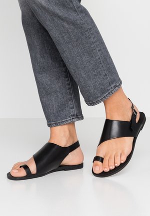 Pieds portant des sandales ouvertes en cuir noir avec des brides à la cheville et un jean gris foncé retroussé sur un fond clair.