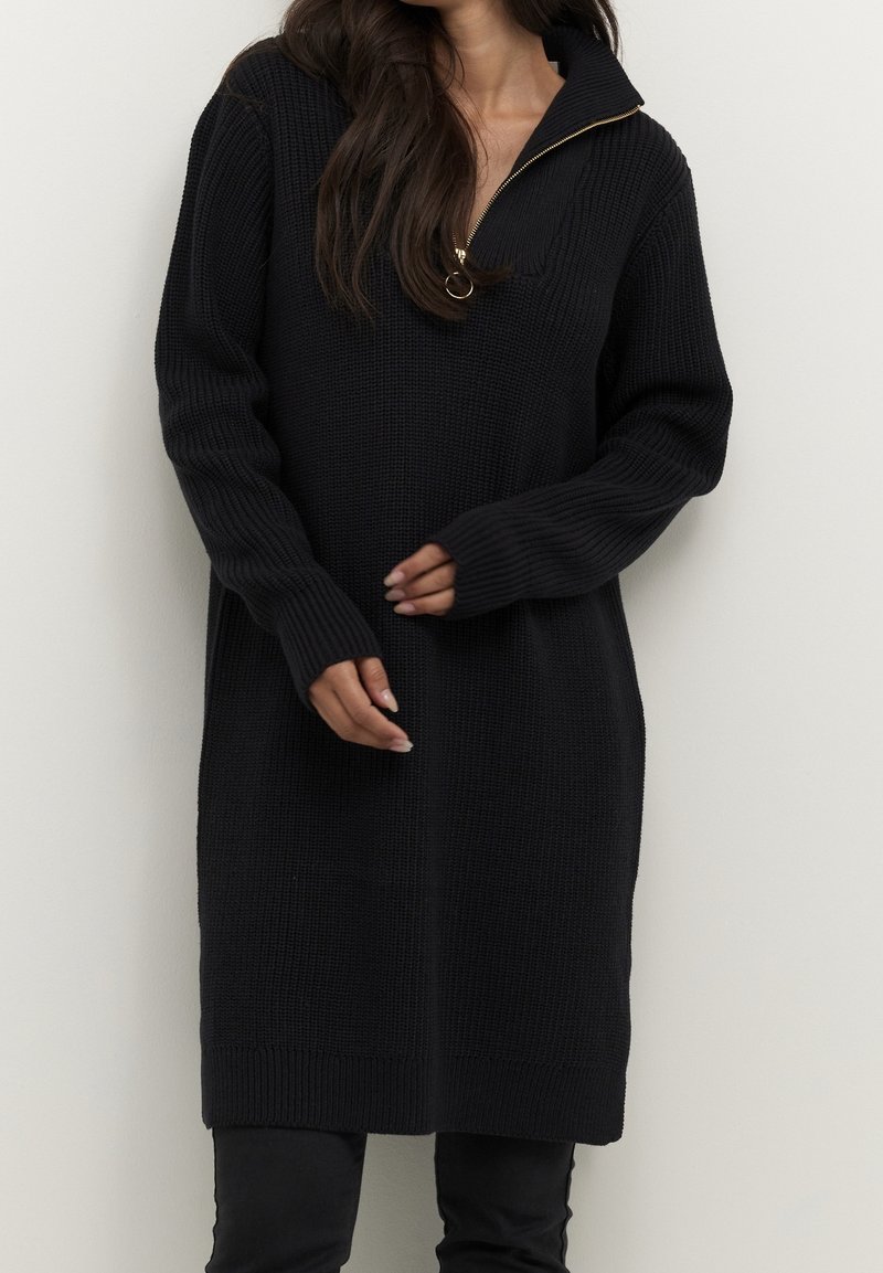 Femme portant un long pull en maille noire avec un col zippé doré partiellement ouvert, debout devant un fond clair uni.