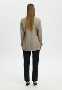 Persona con cabello largo y liso, vistiendo un blazer beige, pantalones negros y tacones negros, de pie de espaldas frente a un fondo liso.