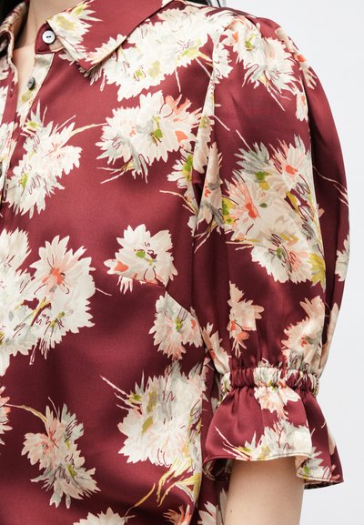 Bordó blúz virágmintával, galléros nyakkal és puffos ujjakkal. Puha anyag, textúrált felülettel és rugalmas mandzsettákkal.