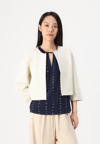 Beige kort kappa med trekvartslånga ärmar, strukturerad tyg, öppen framsida, bärs över en marinblå topp med vita prickar.