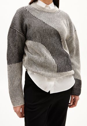 Personne portant un pull en tricot rayé noir et blanc par-dessus une chemise blanche à col et un pantalon noir, debout devant un fond uni.