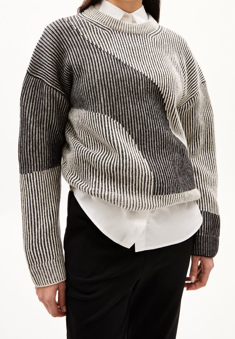 Personne portant un pull en tricot rayé noir et blanc par-dessus une chemise blanche à col et un pantalon noir, debout devant un fond uni.
