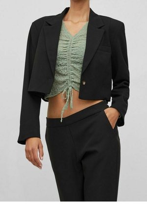 Černý zkrácený blazer s jedním knoflíkem, spárovaný se světle zeleným strukturovaným topem s nařaseným designem a stahovací šňůrkou, doplněný černými kalhotami.