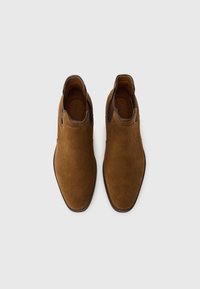 Hnědé semišové Chelsea boty na nazutí s tmavě hnědými elastickými bočními panely a černými podrážkami, pohled shora na bílém pozadí.