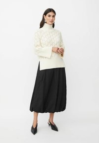Crème gebreid sweater met een hoge halslijn en opengewerkt patroon, gecombineerd met een zwarte volumineuze rok en puntige zwarte slingback schoenen.