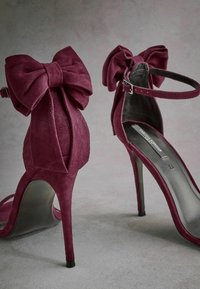 Sandales à talons hauts en suede de couleur bourgogne foncé, ornées de nœuds surdimensionnés au talon, de fines brides de cheville et d'une semelle noire brillante.