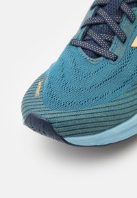 Löpsko med en turkos nätöverdel, texturerad yta och ett kontrasterande marinblå snörsystem. Har en ljusblå mellansula och gummisulor.