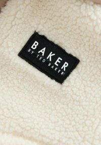 Kreemivärvi fliiskangas pehme tekstuuriga ja must silt tekstiga "BAKER BY TED BAKER", mis on kangasse sisse õmmeldud.