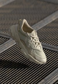 Sneaker beige chunky con tessuto testurizzato e suola spessa poggiato su un pavimento in griglia metallica, illuminato da una luce naturale morbida.