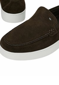 Chaussure slip-on en suède brun foncé avec semelle en caoutchouc blanche et détails cousus, affichant un petit logo métallique sur le dessus.
