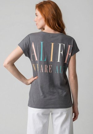 Frau mit roten Haaren, die ein graues T-Shirt mit großem, mehrfarbigem Text "ALIFE" und zusätzlichem kleineren Text trägt, kombiniert mit weißen Hosen.