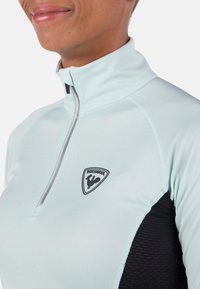 Helles blaues, zip-festes Sportoberteil mit einem schwarzen Strukturelement, hohem Kragen und einem Logo-Patch auf der Brust. Glatter, eng anliegender Stoff.