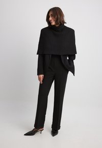 Schwarzer, gerippter Strick-Turtleneck-Cape, getragen über einem figurschmeichelnden schwarzen Anzug und maßgeschneiderten Hosen. Das Outfit wird mit spitz zulaufenden schwarzen Mules kombiniert.