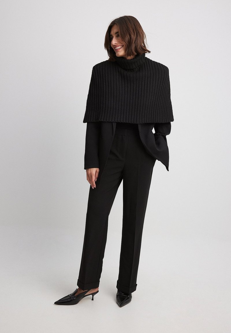 Mustat ribatut neule-turtleneck-viitta, joka on puettu tiukan mustan puvun ja muotoiltujen housujen päälle. Asusteena on teräväkärkiset mustat mules-kengät.