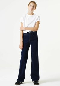 Donkerblauwe recht gesneden spijkerbroek met hoge taille, voorzien van vijf zakken en contrasterende stiksels. Gecombineerd met een wit T-shirt met korte mouwen.