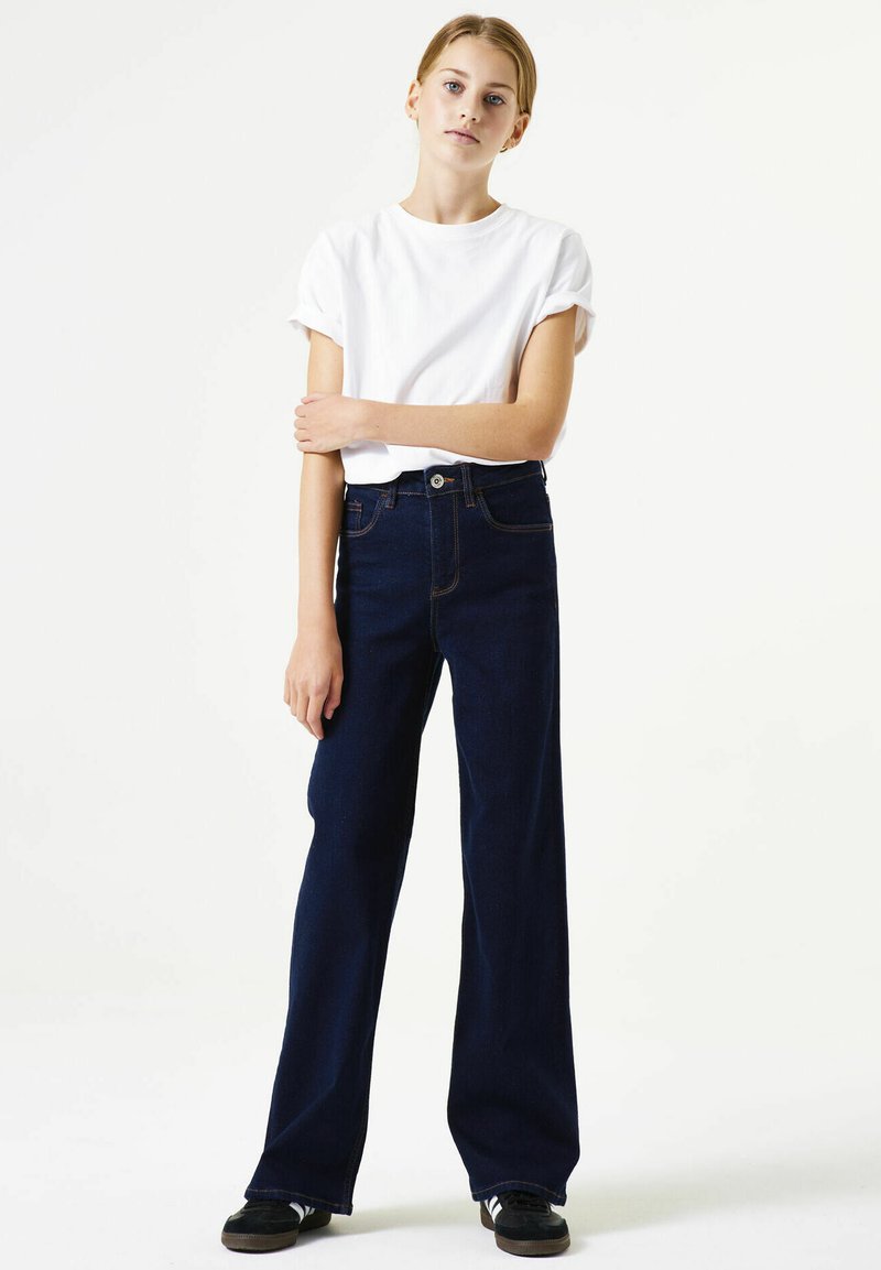 Donkerblauwe recht gesneden spijkerbroek met hoge taille, voorzien van vijf zakken en contrasterende stiksels. Gecombineerd met een wit T-shirt met korte mouwen.